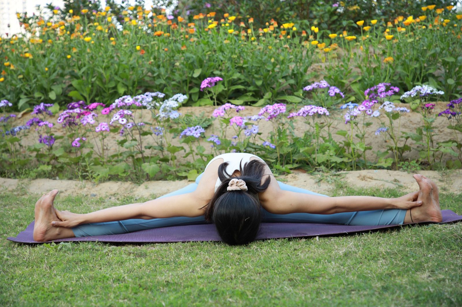 Juhie in a wide-legged seated forward fold on a mat in a flower garden
