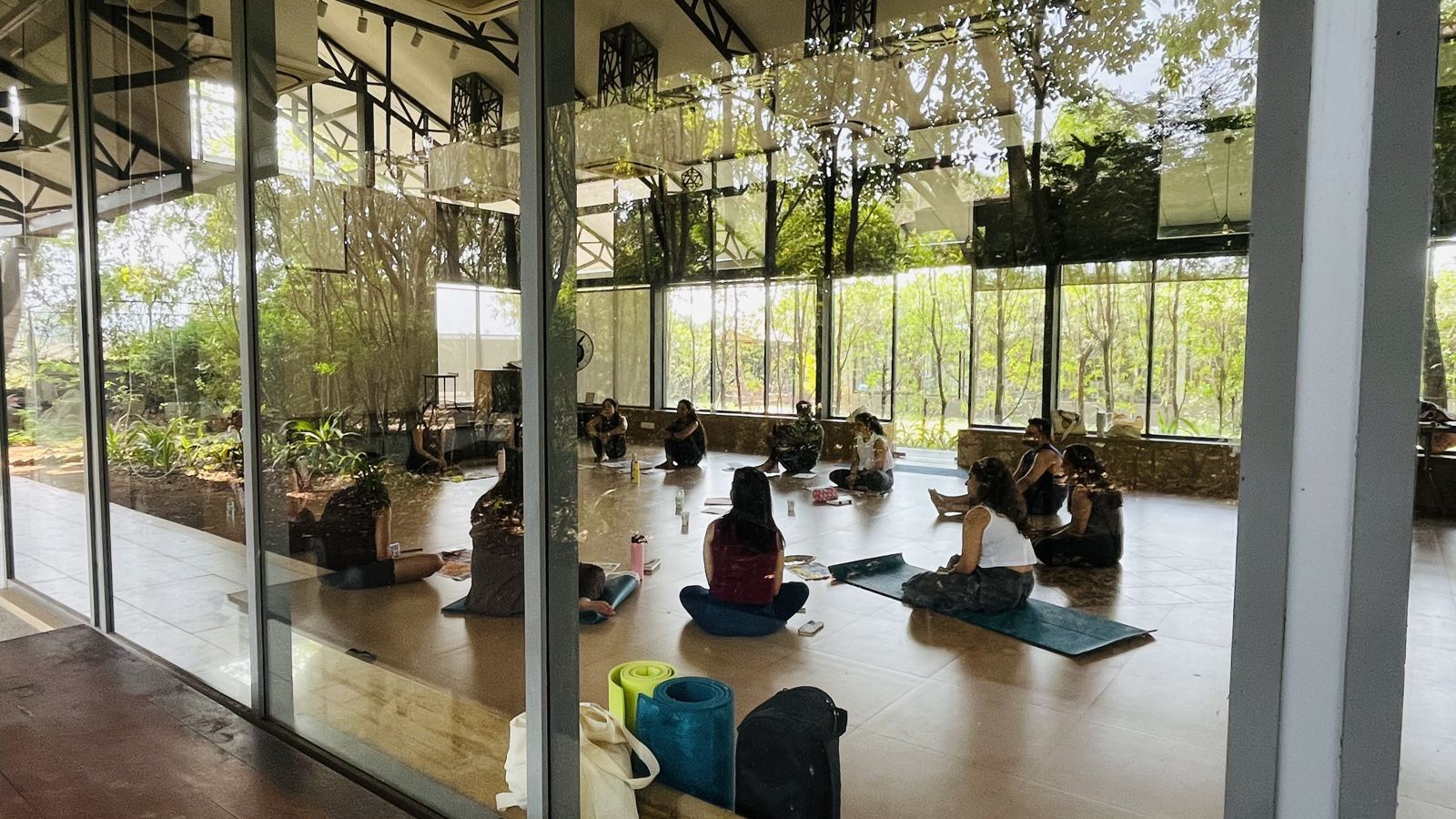A retreat group practising yoga in a glass-walled hall surrounded by trees in Karjat