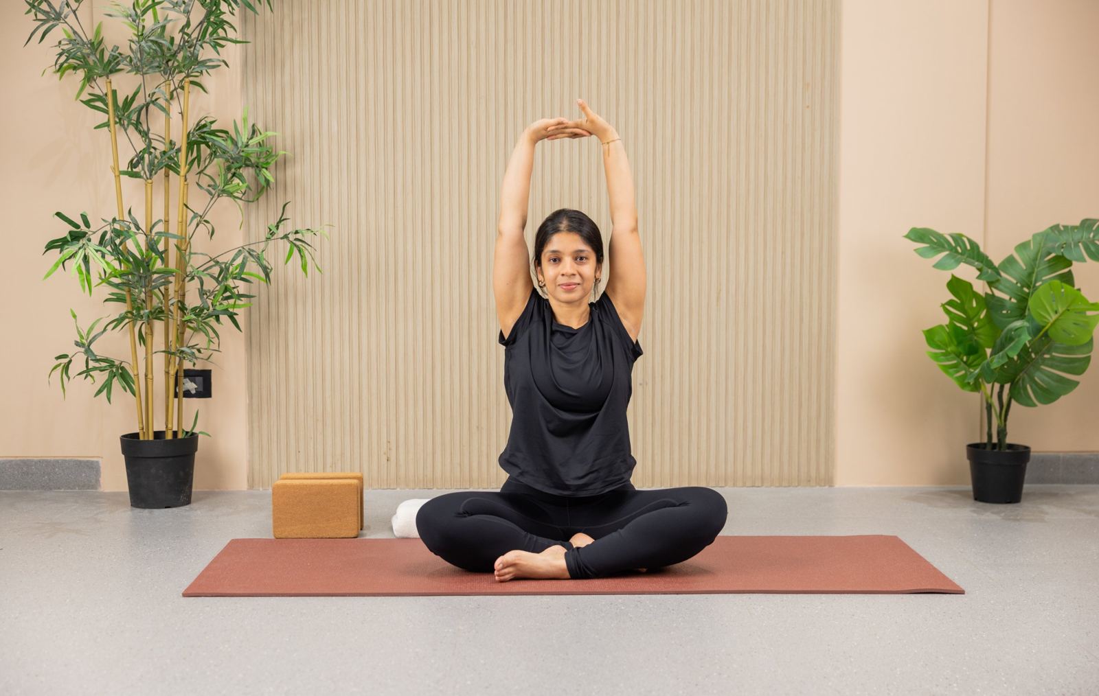 Juhie smiling, seated on a mat with arms stretched overhead in a yoga studio